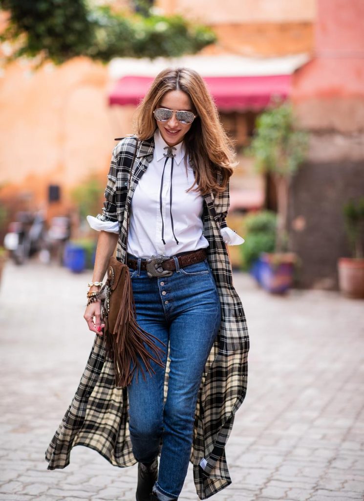Alexandra Lapp in a Cowboy Style Look wearing a long checkered shirt dress from Polo Ralph Lauren, a white shirt from Loro Piana, light blue denims from Adriano Goldschmied, a brown suede leather bag with long fringes and a buckle with feathers from Saint Laurent, a black leather belt with white stitches from Isabel Marant, black Cowboy boots from Christian Dior, a silver Chanel brooch and mirrored sunglasses from Givenchy on November 25, 2018 in Marrakech, Morocco.