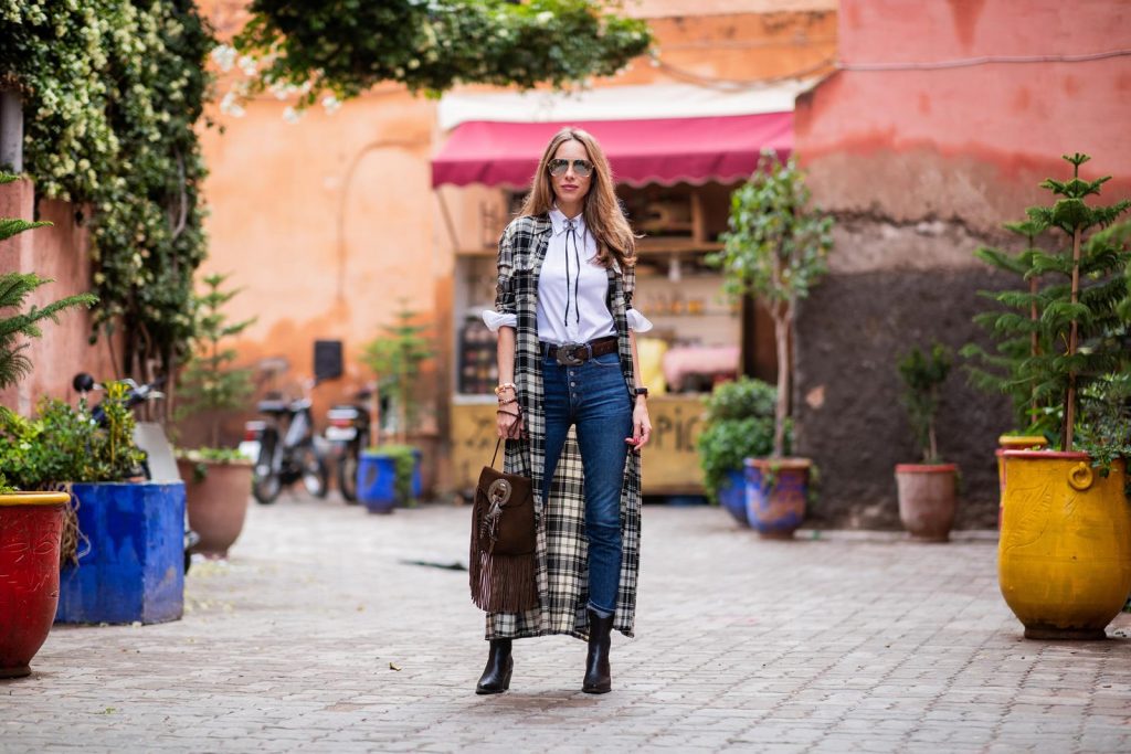 Alexandra Lapp in a Cowboy Style Look wearing a long checkered shirt dress from Polo Ralph Lauren, a white shirt from Loro Piana, light blue denims from Adriano Goldschmied, a brown suede leather bag with long fringes and a buckle with feathers from Saint Laurent, a black leather belt with white stitches from Isabel Marant, black Cowboy boots from Christian Dior, a silver Chanel brooch and mirrored sunglasses from Givenchy on November 25, 2018 in Marrakech, Morocco.